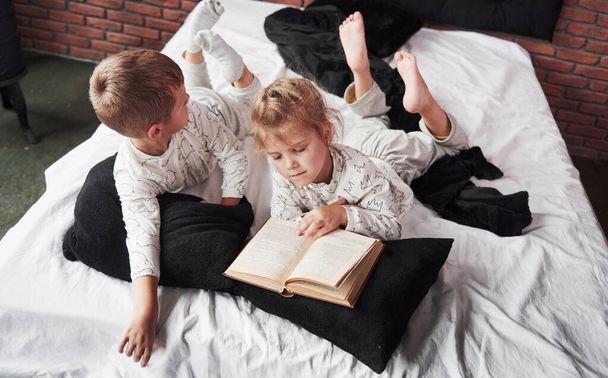 Two children lie on a large bed and read an interesting book. They are dressed in the same pajamas. - Фото, изображение