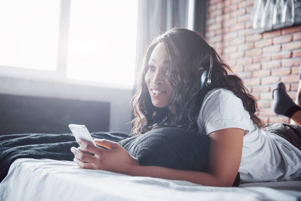Beautiful african girl in sleepwear smiling listening to music in headphones lying on bed. - Фото, изображение