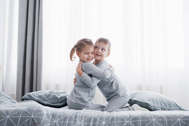 Happy kids playing in white bedroom. Little boy and girl, brother and sister play on the bed wearing pajamas. Nursery interior for children. Nightwear and bedding for baby and toddler. Family at home. - Foto, afbeelding