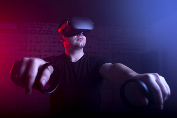 VR gözlüklü bir oyuncu gece oyun odasında bir tetikçiyi oynar, sanal dünyada oyun kumandalarıyla ateş eden bir adam. - Fotoğraf, Görsel