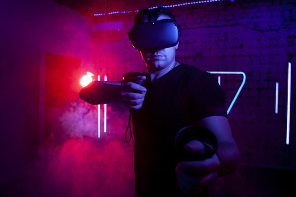 guy a gamer in VR glasses plays a shooter in the game room at night, a man shoots in the virtual world with game joysticks - Photo, Image