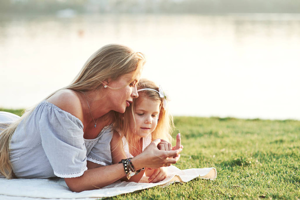 Kid is wondering what sort of nail polish mommy has use. Photo of young mother and her daughter having good time on the green grass with lake at background. - 写真・画像