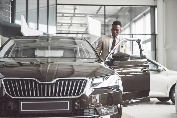 Young black businessman on auto salon background. Car sale and rent concept. Rich african american man. - 写真・画像