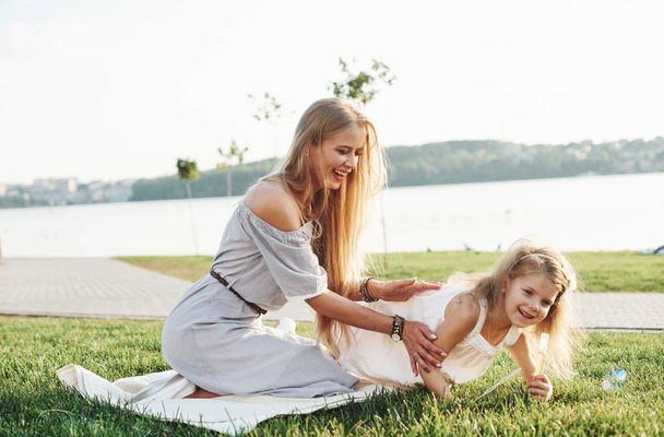 Photo of young mother and her daughter having good time on the green grass with lake at background. - Фото, изображение