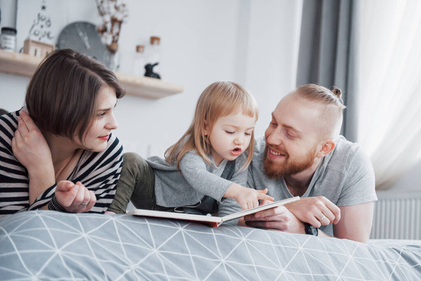 Father, Mother and Little Daughter Reading Children's Book on a Sofa in the Living Room. Happy big family read an interesting book on a festive day. Parents love their children. - Foto, Bild