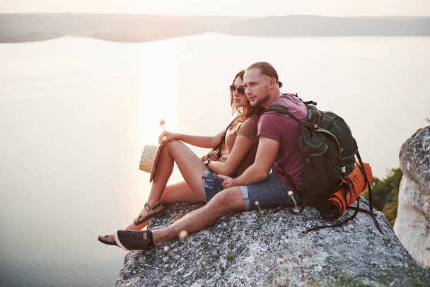 Hugging couple with backpack sitting on top of rock mountain enjoying view coast a river or lake. Traveling mountains and coast, freedom and active lifestyle concept. - Foto, imagen