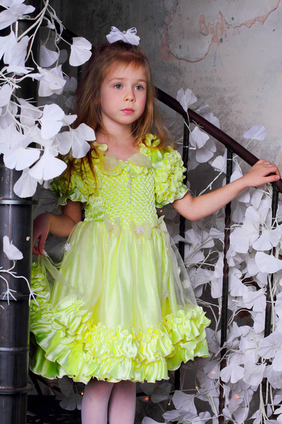a little Princess with an angelic face in a yellow dress stands on the stairs among the white flowers - Photo, Image