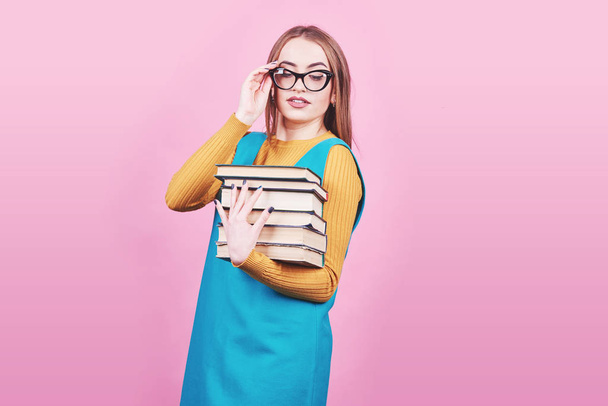 Happy cute girl in glasses holding in hands a pile of books isolated on colorful pink background. - Фото, зображення