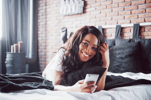Portrait of beautiful woman waking up in her bed and looks into the phone. Check social networks, send sms. The girl is wearing a T-shirt. - Fotografie, Obrázek