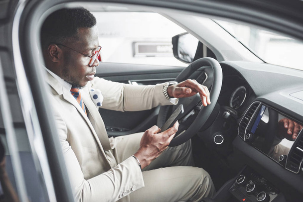 Stylish black businessman sitting behind the wheel of new luxury car. Rich african american man using smartphone. - Photo, image