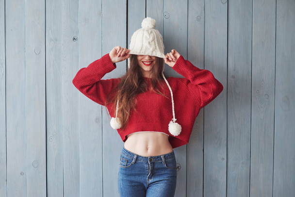 How long this thing is. It's covering my eyes. Funny girl in knitted sweater and hat standing indoors with wooden background. - Фото, изображение