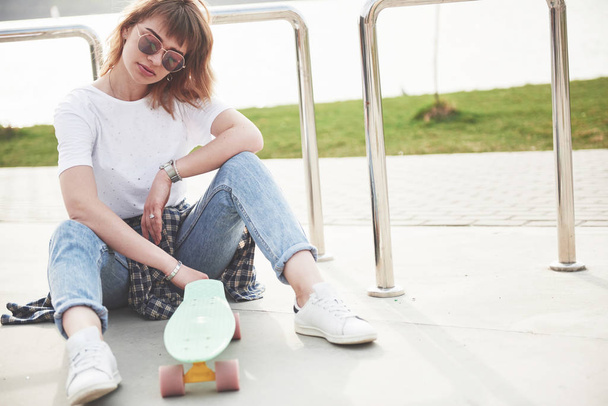 ein Foto eines schönen Mädchens mit schönen Haaren hält ein Skateboard auf einem langen Brett, blickt in die Kamera und lächelt, urbanes Leben - Foto, Bild