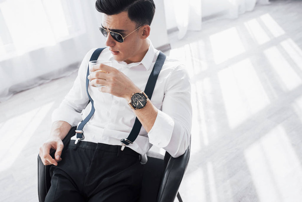 Fashion concept. Young stylish businessman sitting on a black chair with glass of whiskey. - Фото, изображение