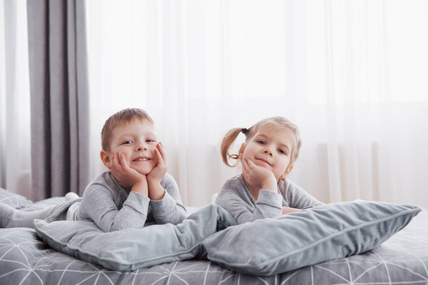 Happy kids playing in white bedroom. Little boy and girl, brother and sister play on the bed wearing pajamas. Nightwear and bedding for baby and toddler. Family at home. - Foto, Bild