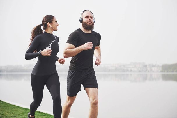 Couple jogging and running outdoors in park near the water. Young bearded man and woman exercising together in morning. - Φωτογραφία, εικόνα