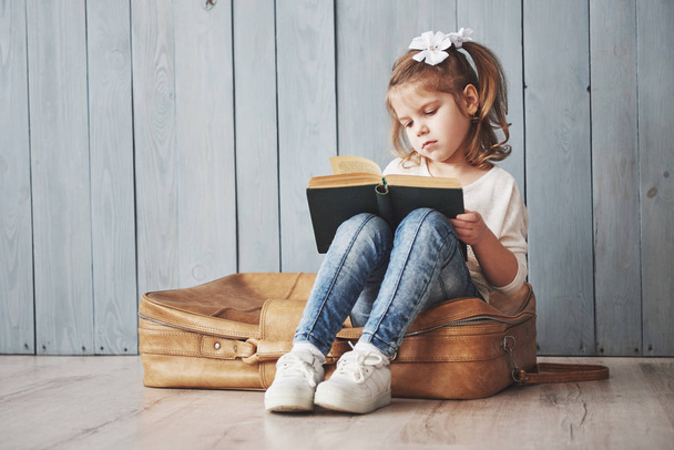 Ready to big travel. Happy little girl reading interesting book carrying a big briefcase. Freedom and imagination concept. - 写真・画像