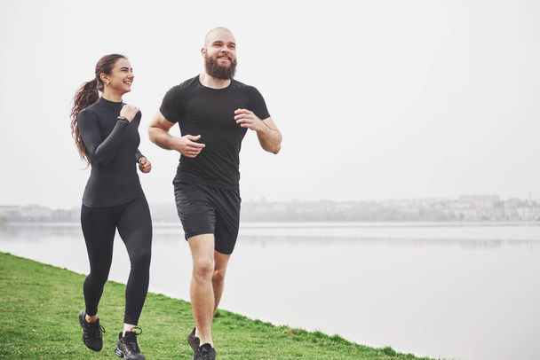 Couple jogging and running outdoors in park near the water. Young bearded man and woman exercising together in morning. - Fotoğraf, Görsel