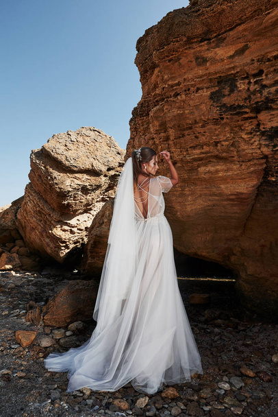 Portrait of a girl in a wedding gorgeous dress posing a photographer on the beach. The bride is on the rocks. - Foto, Imagen