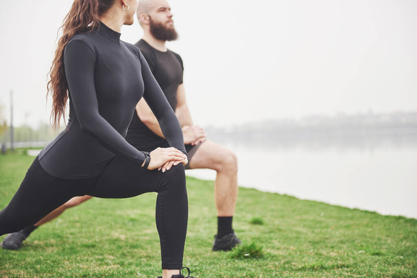 Fitness couple stretching outdoors in park near the water. Young bearded man and woman exercising together in morning. - Foto, imagen
