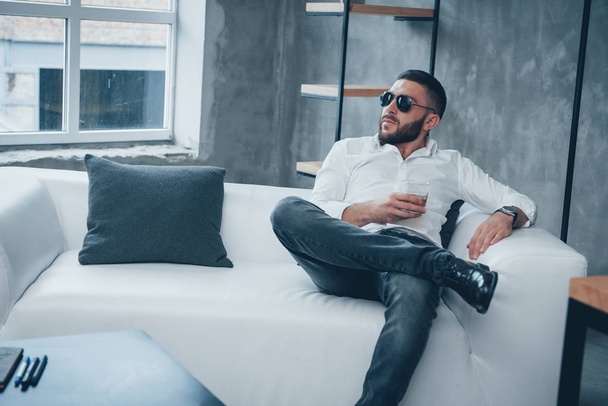 Thoughtful serious look. Young short-haired man in sunglasses sitting on couch in the office. - Foto, Bild