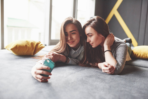 Two cute smiling twins sisters holding smartphone and making selfie. Girls lie on the couch posing and joy. - Foto, Imagen