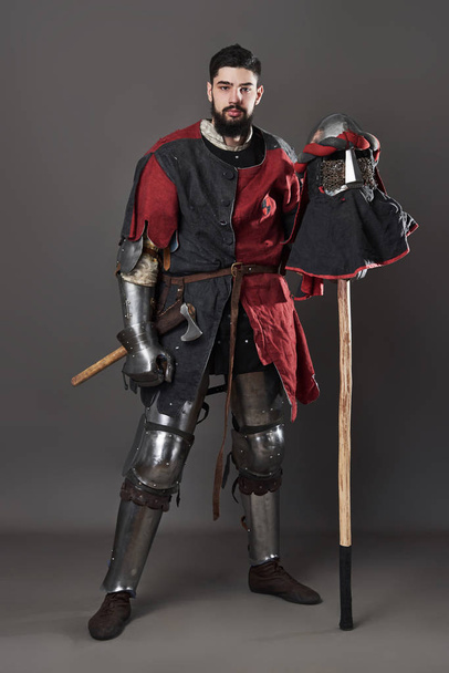 Medieval knight on grey background. Portrait of brutal dirty face warrior with chain mail armour red and black clothes and battle axe. - Foto, afbeelding