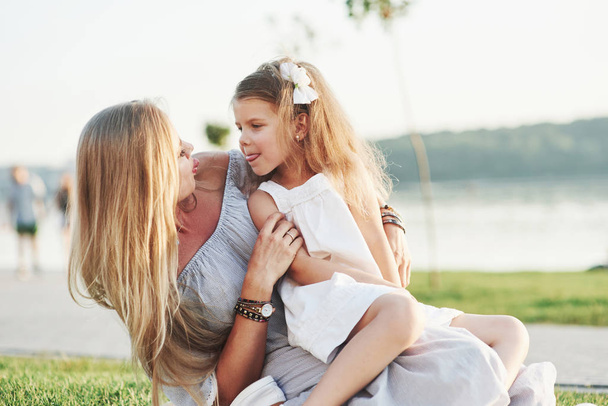 Teasing. Photo of young mother and her daughter having good time on the green grass with lake at background. - 写真・画像