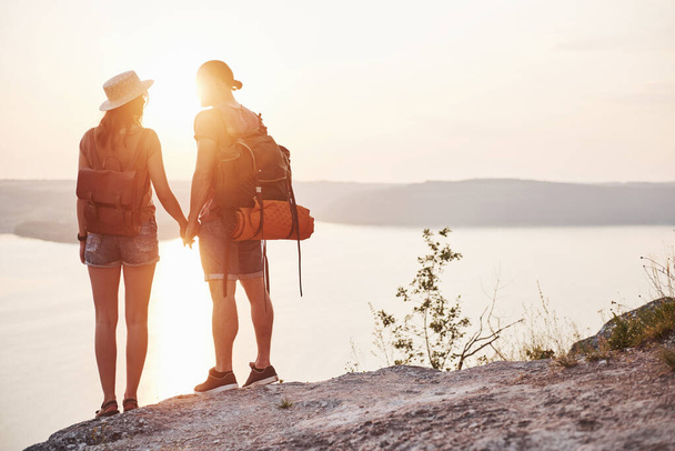 Two tourist male and woman with backpacks stand to the top of the crag and enjoying sunrise. Traveling mountains and coast, freedom and active lifestyle concept. - Fotó, kép