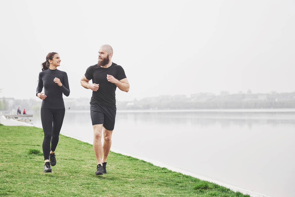 Couple jogging and running outdoors in park near the water. Young bearded man and woman exercising together in morning. - Valokuva, kuva