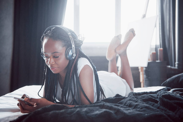 Beautiful african girl in sleepwear smiling listening to music in headphones lying on bed. - Foto, afbeelding