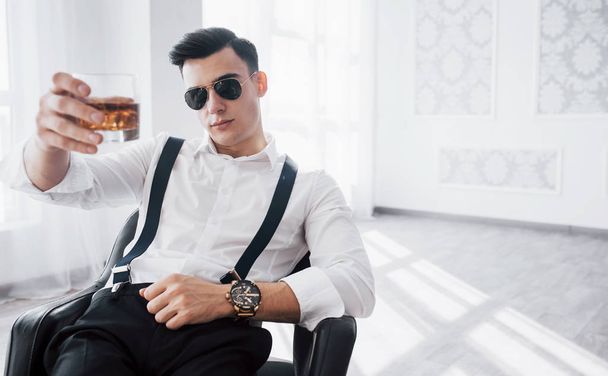 Let's take a shot. Young stylish businessman in sunglasses sitting on a black chair with glass of whiskey. - Fotó, kép
