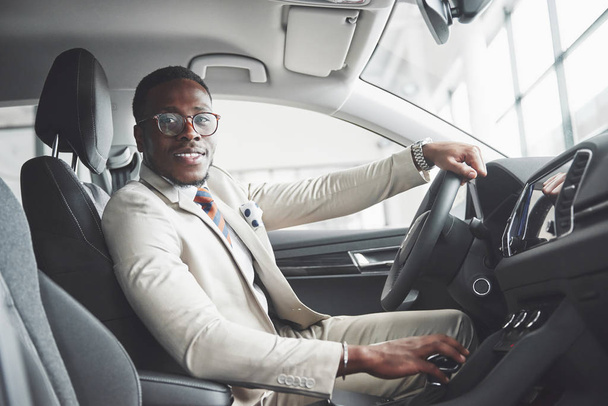 Stylish black businessman sitting behind the wheel of new luxury car. Rich african american man using smartphone. - Zdjęcie, obraz