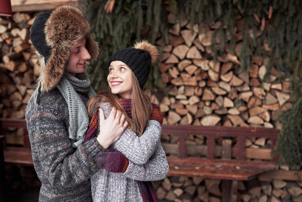 winter, vacation, couple, christmas and people concept - smiling man and woman in hats and scarf hugging over wooden country house and snow background. - Φωτογραφία, εικόνα