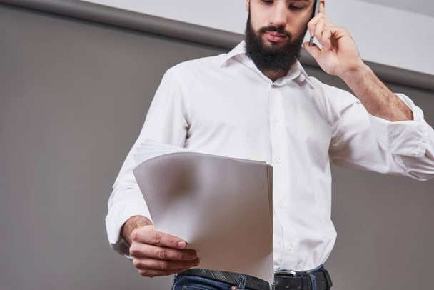 Business man with beard in white shirt holding documents and phone on gray background. - Φωτογραφία, εικόνα