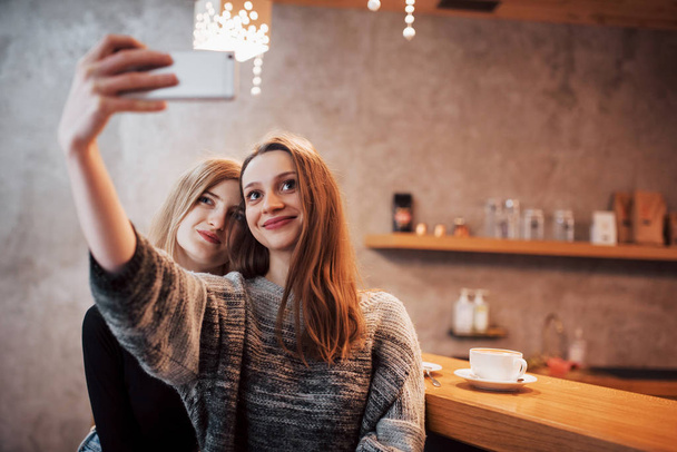 Двоє друзів п'ють каву в кафе, приймають селфі зі смартфоном і розважаються, роблячи смішні обличчя. Фокус на дівчині зліва
. - Фото, зображення