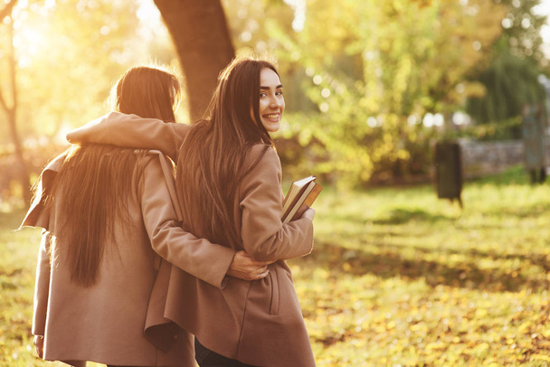 View from the back of young smiling brunette twin girls hugging and having fun in casual coat walking at autumn sunny park on blurry background. One of them is looking backwards and holding books. - Фото, зображення