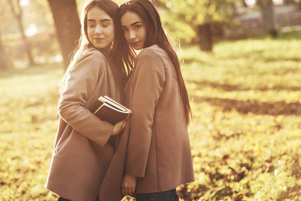 Young charming brunette twin girls standing very close turned to each other in casual coat , touching heads at autumn sunny park on blurry background. One of them is holding books with eyes closed. - Foto, Imagem