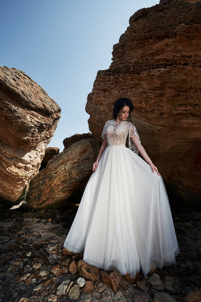 Portrait of a girl in a wedding gorgeous dress posing a photographer on the beach. The bride is on the rocks. - Φωτογραφία, εικόνα