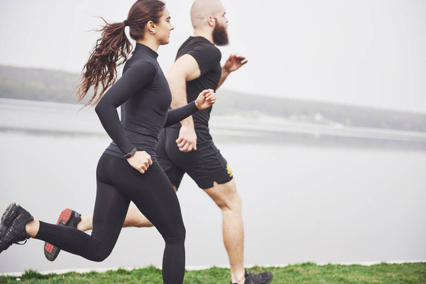 Couple jogging and running outdoors in park near the water. Young bearded man and woman exercising together in morning. - Photo, image