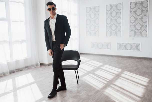 Spacious almost empty hall with black chair. Portrait of young stylish man in classic business wear standing in the white room. - Foto, Imagen
