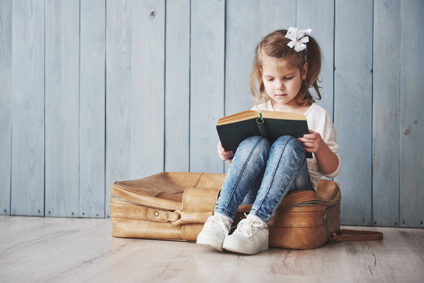 Ready to big travel. Happy little girl reading interesting book carrying a big briefcase and smiling. Travel, freedom and imagination concept. - Foto, Imagen