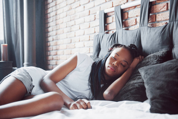 A beautiful African woman lies and sleeps in bed, in a white bedroom. Good morning, vacation at home. The girl is wearing a T-shirt, pigtails on her head. - Foto, Imagem