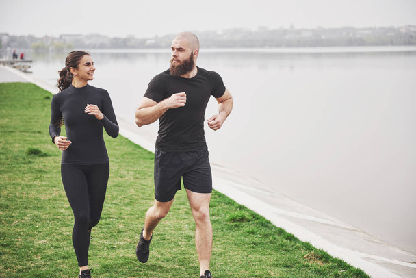 Couple jogging and running outdoors in park near the water. Young bearded man and woman exercising together in morning. - Photo, Image