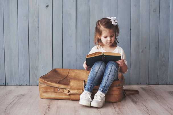 Ready to big travel. Happy little girl reading interesting book carrying a big briefcase and smiling. Travel, freedom and imagination concept. - 写真・画像