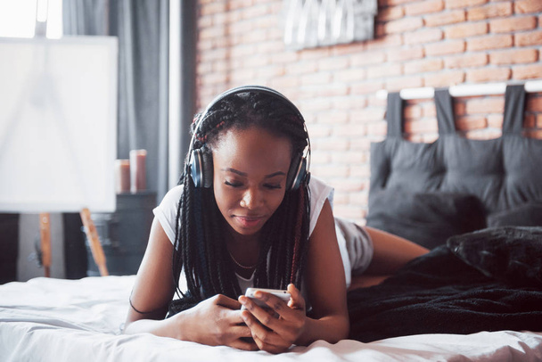 Hermosa chica africana en ropa de dormir sonriendo escuchando música en auriculares acostados en la cama
. - Foto, Imagen
