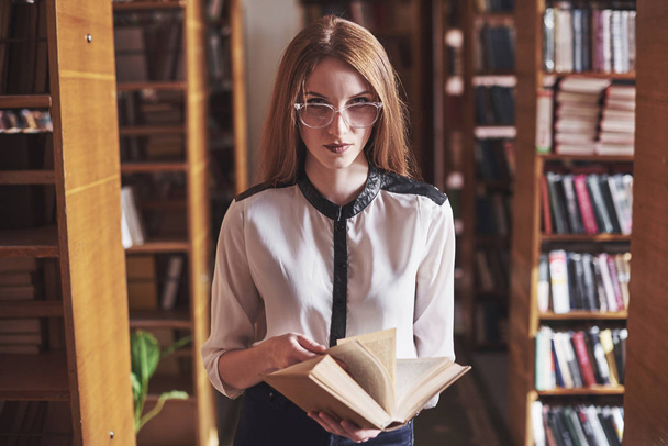 Young attractive student librarian reading a book between library bookshelves. - Fotó, kép