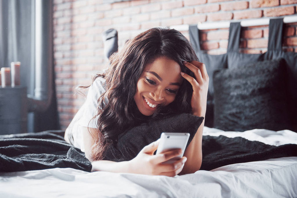 Portrait of beautiful woman waking up in her bed and looks into the phone. Check social networks, send sms. The girl is wearing a T-shirt. - Photo, Image