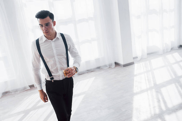 Empty room. Guy in the elegant luxury suit walking in the spacious white room with whiskey in the hand. - Foto, imagen