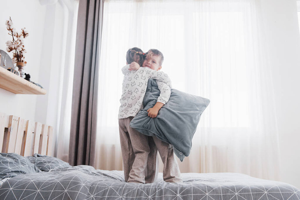Happy kids playing in white bedroom. Little boy and girl, brother and sister play on the bed wearing pajamas. Nursery interior for children. Nightwear and bedding for baby and toddler. Family at home - 写真・画像