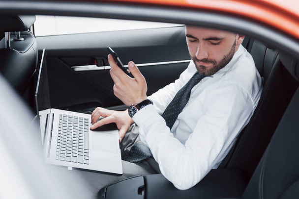 A young businessman working on laptop and talking on the phone while sitting in the car's back. Works in motion, appreciates its time. - Foto, Bild